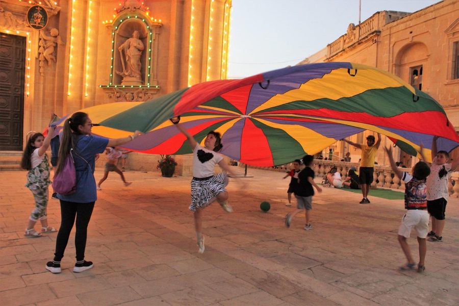 Children and volunteers playing with a large rainbow parachute in a historic town square at dusk